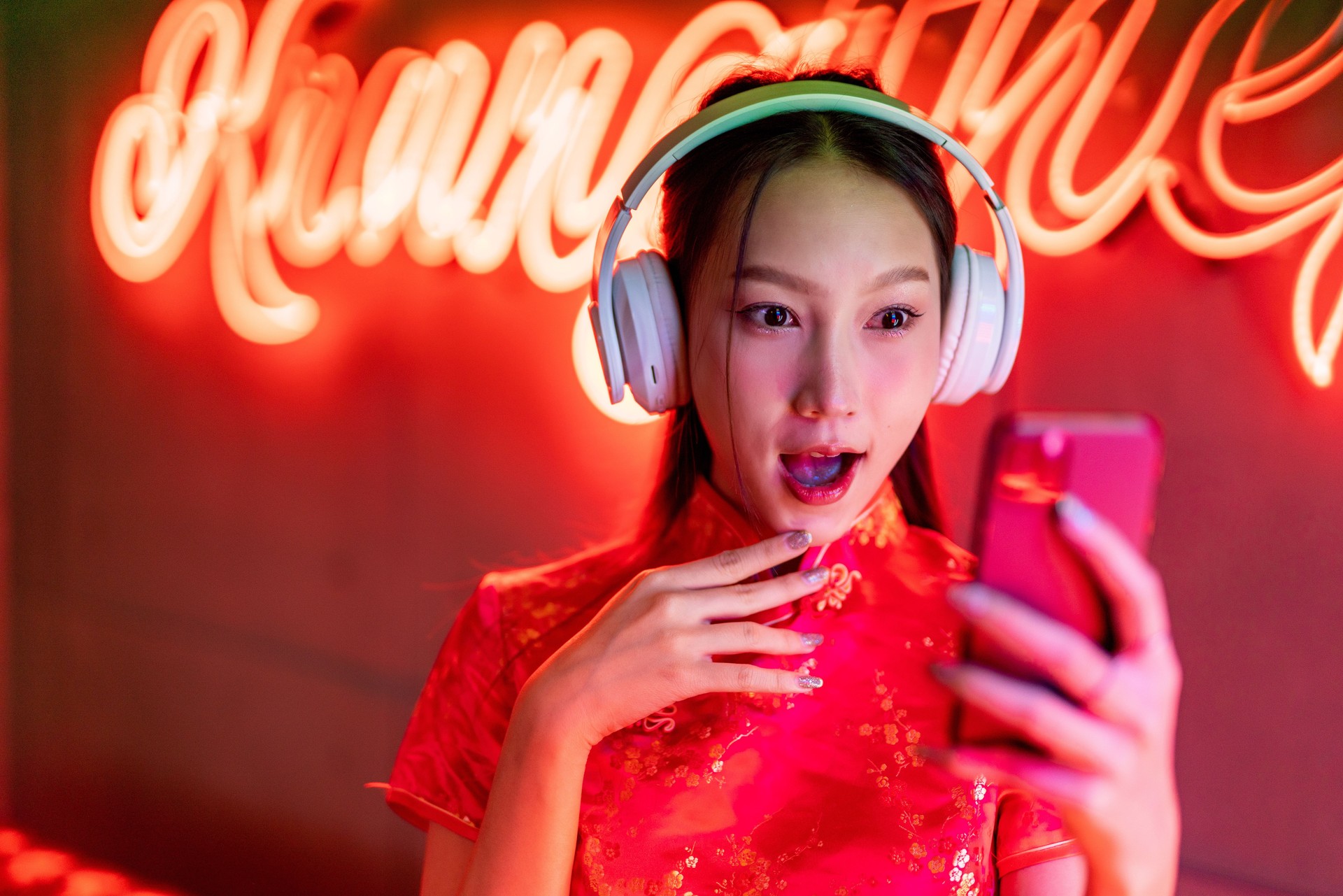 Portrait photo of a beautiful young asian female lady model with cute look wearing headphone and red traditional chinese cheongsam dress listening music using her mobile smart phone in front of orange pink neon lights