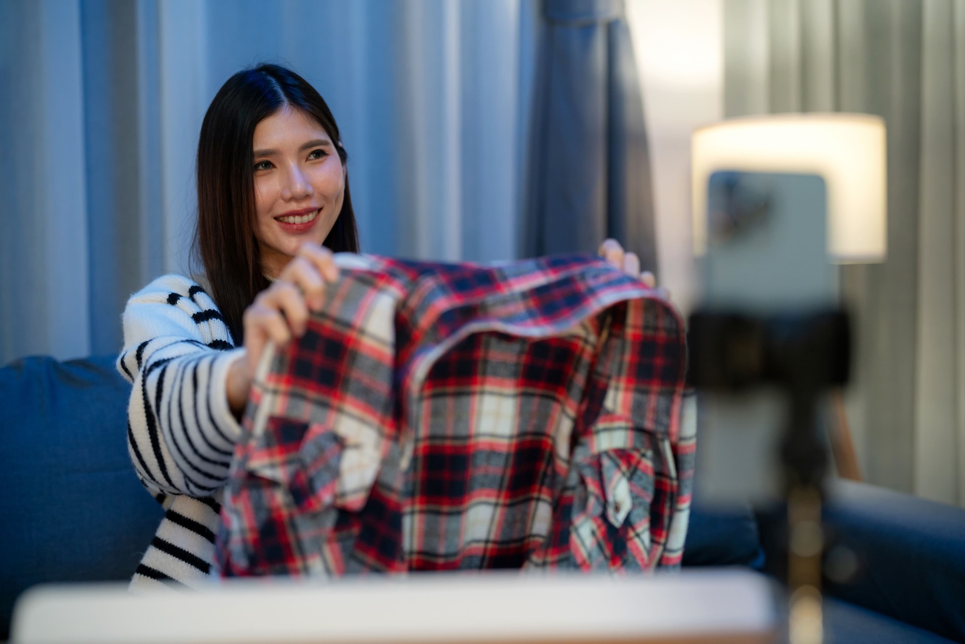 Asian female influencer livestreaming from her cozy living room, showcasing trendy women's clothing via an online platform. A perfect blend of social media marketing, home shopping, and digital entrepreneurship with real-time customer engagement.